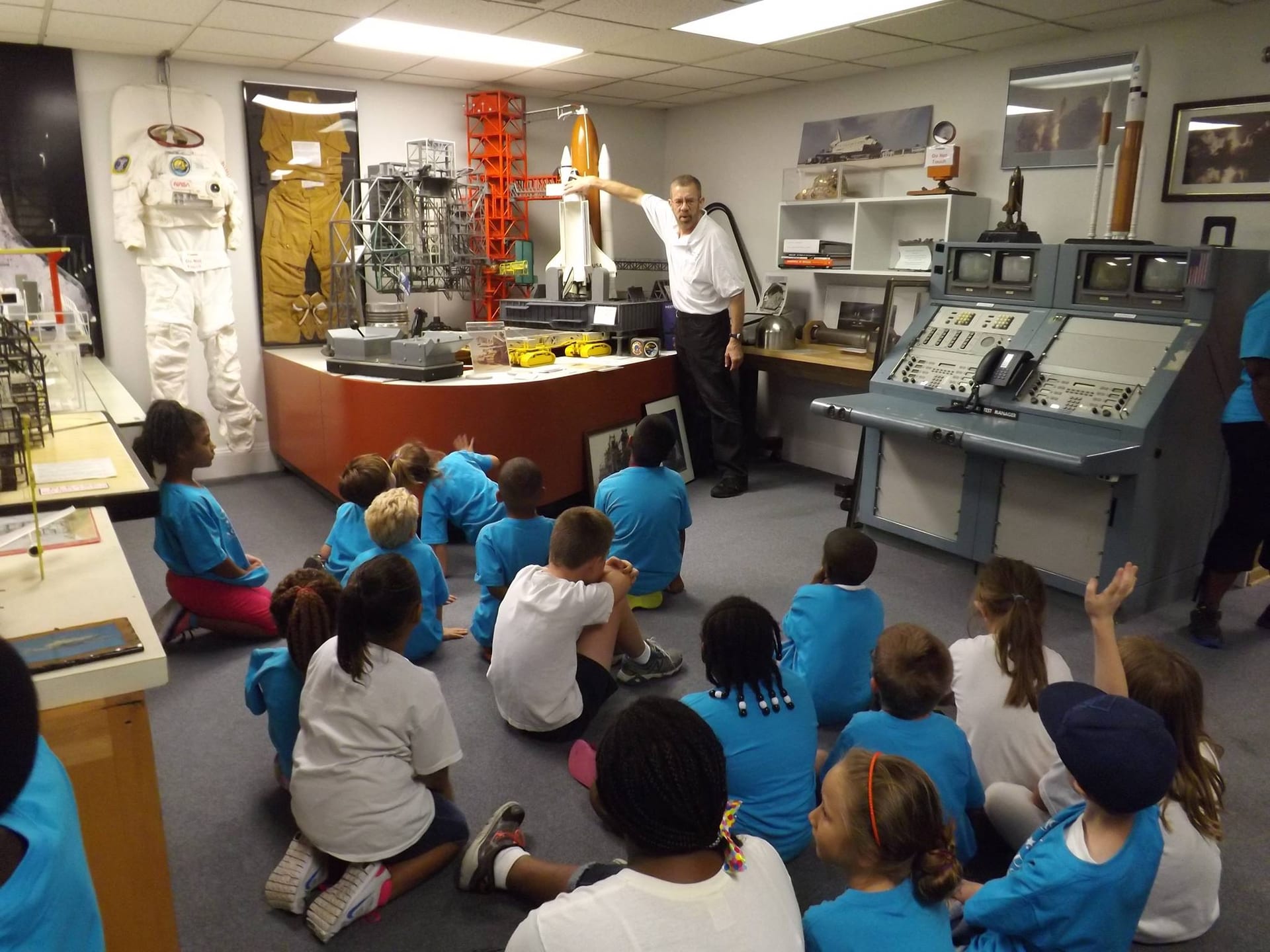 A museum docent addresses seated students in a gallery of shuttle launch consoles.